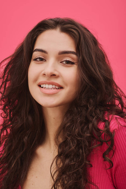 Primer plano de la joven con el pelo largo ondulado sonriendo a la cámara aislada sobre fondo rosa - Foto, Imagen