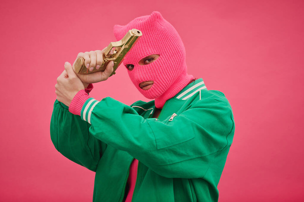 Portrait of bandit woman in pink balaclava crossed her arms with gun in front of her face and looking at camera against pink background - Foto, Imagem