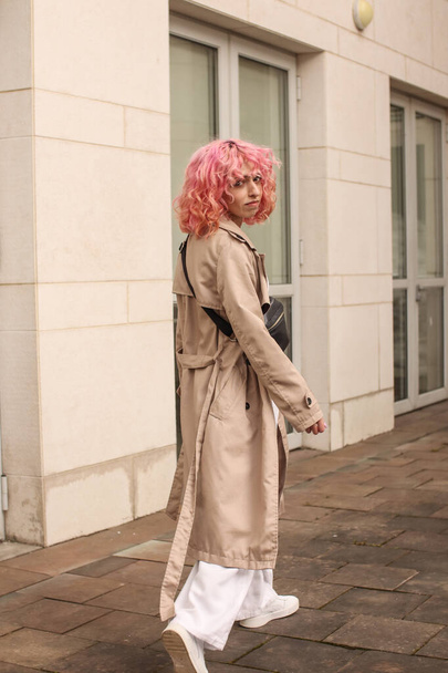 Πορτρέτο γυρίσματα ενός κομψού κοριτσιού με ροζ μαλλιά. Μπεζ αποχρώσεις. Τάσεις της άνοιξης και του καλοκαιριού 2023.Μπεζ Trench μανδύα με λευκό top και παντελόνι. Μαύρη μικρή τσάντα ζώνης - Φωτογραφία, εικόνα