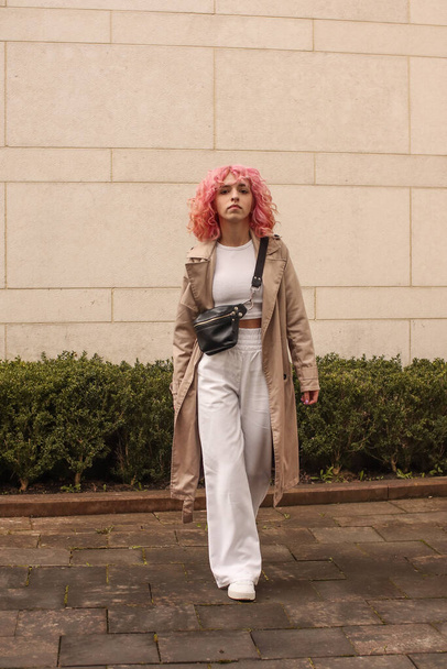 Portrait shooting of a stylish girl with pink hair. Beige shades. Trends of spring and summer 2023.Beige Trench cloak with white top and pants. Black small belt bag - Fotó, kép
