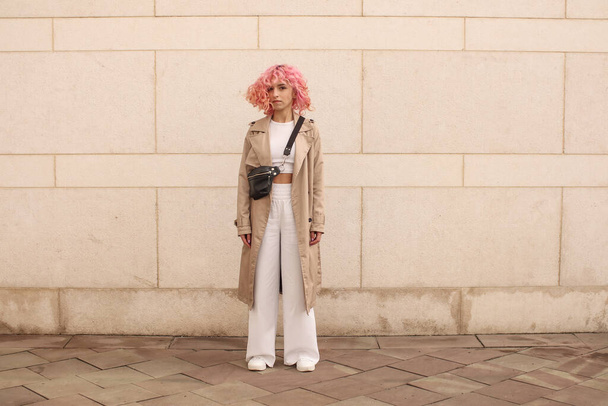 ピンクの髪をしたスタイリッシュな女の子のポートレート撮影。ベージュの色合い。春と夏の傾向2023.ベージュトレンチクローク白トップとパンツと。黒小さなベルトバッグ - 写真・画像