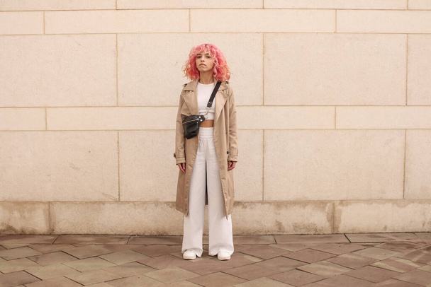 Portrait shooting of a stylish girl with pink hair. Beige shades. Trends of spring and summer 2023.Beige Trench cloak with white top and pants. Black small belt bag - Fotoğraf, Görsel