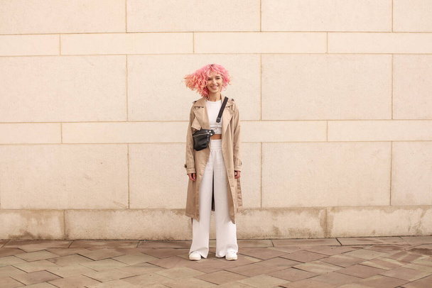 Portrét natáčení stylové dívky s růžovými vlasy. Béžové odstíny. Trendy jara a léta 2023.Béžový plášť s bílou střechou a kalhotami. Černá taška na pásek - Fotografie, Obrázek