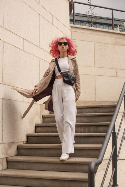 Portrait shooting of a stylish girl with pink hair. Beige shades. Trends of spring and summer 2023.Beige Trench cloak with white top and pants. Black small belt bag - Fotografie, Obrázek