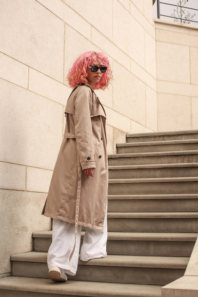 Portrait shooting of a stylish girl with pink hair. Beige shades. Trends of spring and summer 2023.Beige Trench cloak with white top and pants. Black small belt bag - Foto, imagen