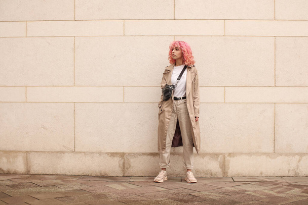 Portrait shooting of a stylish girl with pink hair. Beige shades. Trends of spring and summer 2023. Beige trench cloak with white t-shirt and beige pants. Black small belt bag - Foto, afbeelding