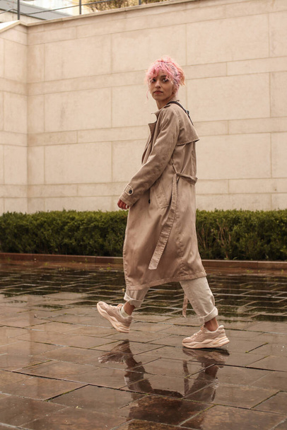 Portrait shooting of a stylish girl with pink hair. Beige shades. Trends of spring and summer 2023. Beige trench cloak with white t-shirt and beige pants. Black small belt bag - Foto, imagen