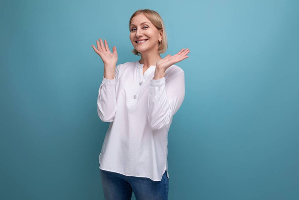 χαρούμενη ξανθιά 50άρα γυναίκα με λευκή μπλούζα στην εμμηνόπαυση στο φόντο του στούντιο. - Φωτογραφία, εικόνα