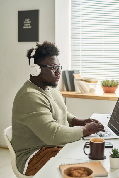 Desarrollador de software joven africano en auriculares inalámbricos que se concentran en su trabajo en línea en el ordenador portátil - Foto, Imagen