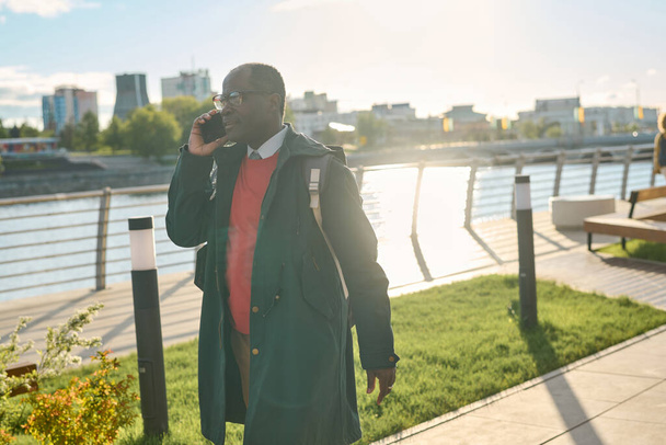 Africano maduro hombre de negocios hablando por teléfono móvil mientras camina por la calle urbana durante el día soleado - Foto, imagen