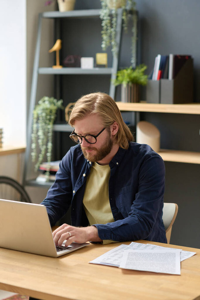 Νεαρός επιχειρηματίας με γυαλιά ηλίου δακτυλογραφεί σε φορητό υπολογιστή και εργάζεται στο διαδίκτυο στο χώρο εργασίας του στο γραφείο - Φωτογραφία, εικόνα