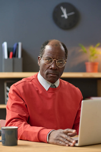 彼の職場にノートパソコンを持って座って、真剣な表情でカメラを見ている眼鏡でアフリカの成熟したビジネスマンの肖像画 - 写真・画像
