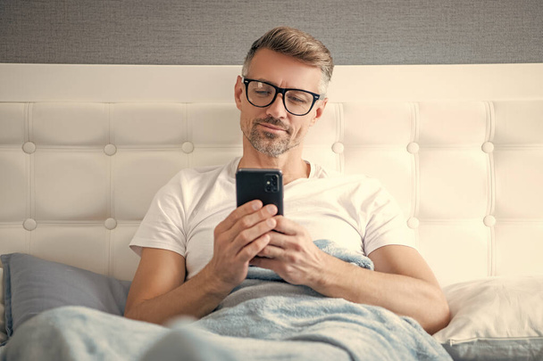 cheerful mature man in bed chatting on phone wearing eyeglasses. - Valokuva, kuva