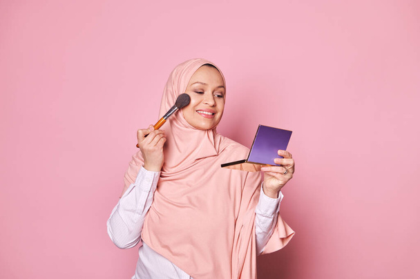 Attraente donna araba musulmana di mezza età in hijab rosa, che tiene la tavolozza con prodotti cosmetici, utilizzando pennello applicando trucco blush sul suo viso, sorridendo un sorriso dentato, isolato su sfondo di colore rosa - Foto, immagini
