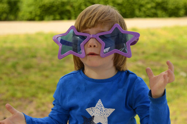 Cute kid with long hair wearing funny star glasses. The boy is preparing for the carnival in the park. - Фото, зображення