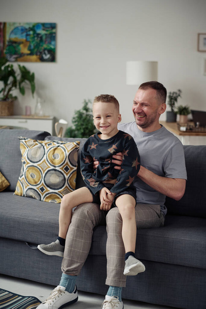Szczęśliwy mały chłopiec siedzi na kolanach swojego taty i bawi się z nim na kanapie w salonie - Zdjęcie, obraz