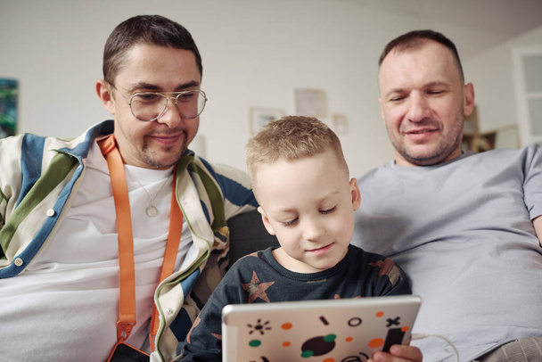 Pequeño niño viendo vídeo en la tableta digital junto con sus padres homosexuales durante el tiempo libre en casa - Foto, imagen