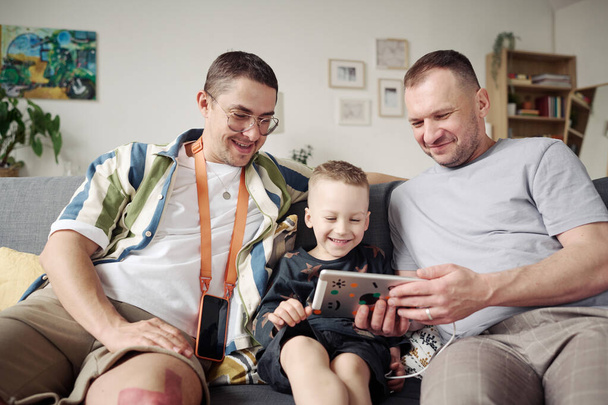 gej para siedzi na kanapie z ich adoptowany syn i oglądać śmieszne wideo na cyfrowy tablet - Zdjęcie, obraz