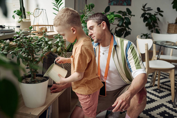 Kleine zoon drenkt groene planten met gieter terwijl vader hem leert om voor ze te zorgen - Foto, afbeelding