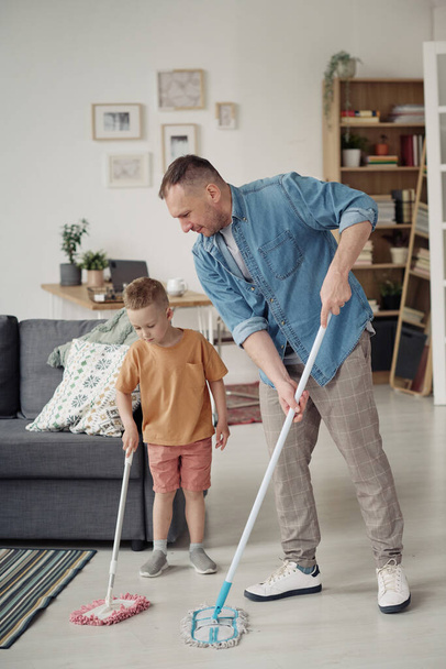 Gay father teaching his adopted child to wash floor with mop during housework in the room - Foto, Bild