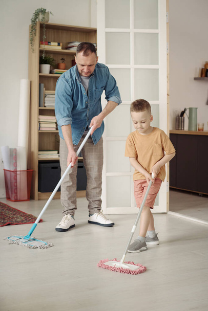 Pequeño hijo lavando el piso con fregona junto con su padre, él le ayuda con las tareas en casa - Foto, imagen