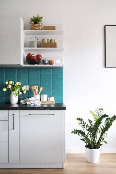 detail in de keuken interieur, witte kast met donker marmeren aanrecht, blauwe tegel op de muur, keramische theepot en kopjes op de plank en potten kamerplant op de vloer - Foto, afbeelding