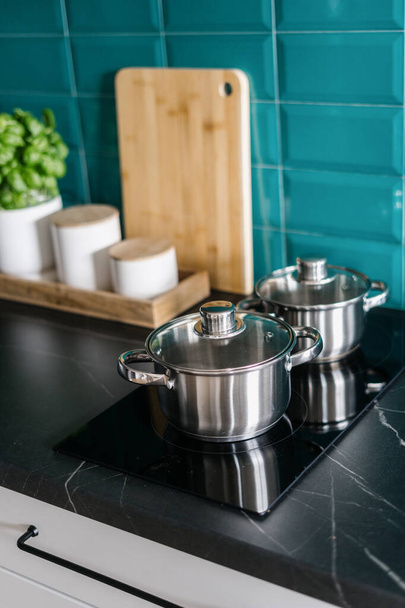 Nahaufnahme von Edelstahltöpfen mit Deckeln auf elektrischem Glaskeramikherd in moderner Küche mit Arbeitsplatte aus schwarzem Marmor und blauer Kachel an der Wand - Foto, Bild