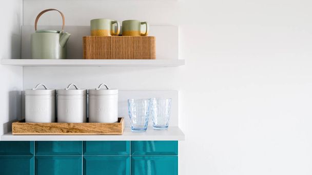 detail at kitchen interior, ceramic teapot, clean cups, glasses and metal storage jars with lids on wooden tray on shelf hanging on white wall, organized space concept - Photo, Image