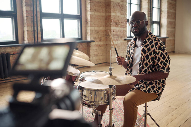 Porträt eines jungen afrikanischen Schlagzeugers, der vor laufender Kamera in einem leeren Studio am Schlagzeug performt - Foto, Bild