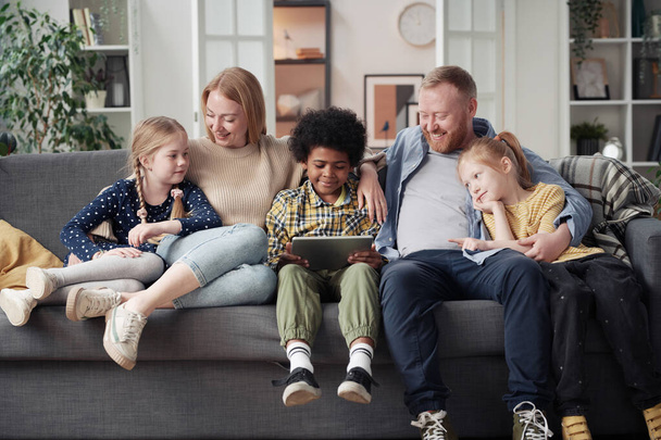 Adoptiolapset katsomassa piirrettyjä digitaalisella tabletilla ja nauttimassa ajasta kasvattivanhempien kanssa sohvalla kotona - Valokuva, kuva