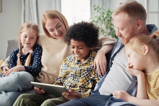 Ευτυχισμένα υιοθετημένα παιδιά βλέποντας βίντεο στο ψηφιακό tablet μαζί με τους θετούς γονείς τους κατά τη διάρκεια του ελεύθερου χρόνου στον καναπέ στο σπίτι - Φωτογραφία, εικόνα