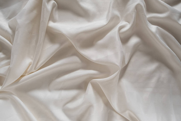 Satin crumpled fabric of light milky color, top view. Natural bed linen, sheets, abstract background of luxury fabric, wavy folds. - Photo, image