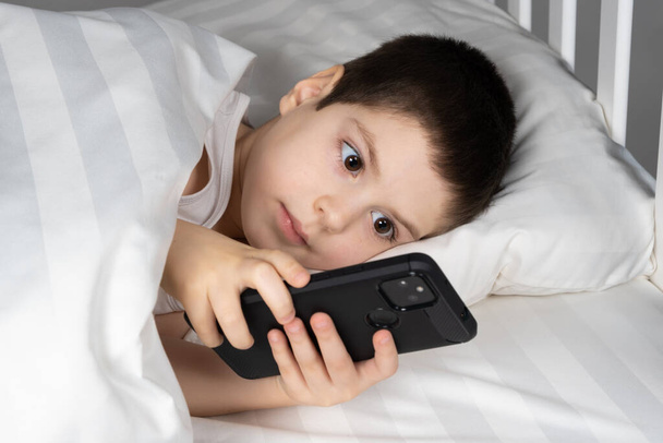 Un garçon de 5 ans joue à des jeux sur son téléphone tout en étant couché dans son lit. Gadgets au lit avant l'heure du coucher chez un enfant. - Photo, image