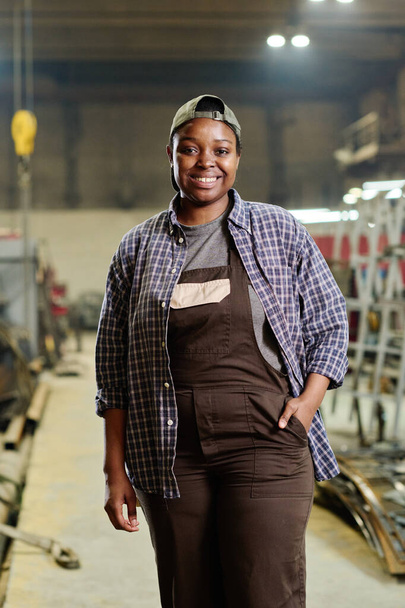 Portrét africké pracovnice v pracovním oděvu usmívající se na kameru stojící v dílně závodu - Fotografie, Obrázek