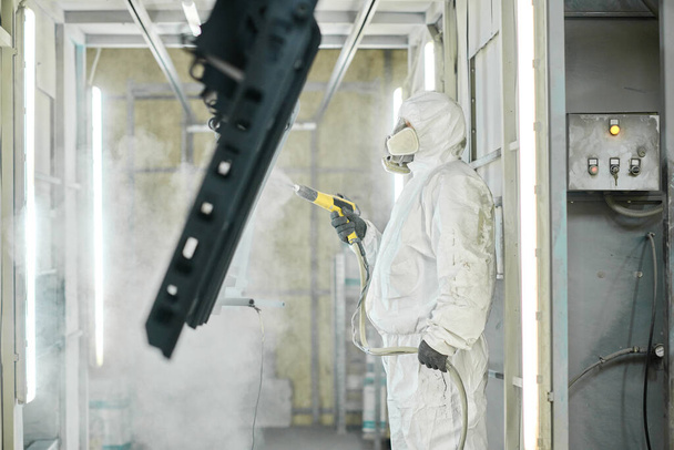 Trabalhador em terno protetor e máscara usando pistola de pulverização para pintar detalhes de aço durante seu trabalho na planta - Foto, Imagem