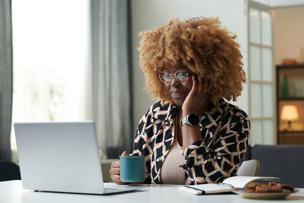 Mujer joven africana con el pelo rizado sentado en la mesa en casa con una taza de café y tener una reunión en línea en el ordenador portátil - Foto, imagen