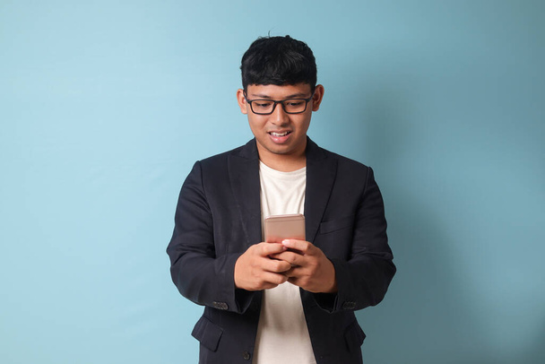 Портрет молодого азиатского бизнесмена в повседневном костюме с широкой улыбкой, держащего телефон обеими руками. Изолированное изображение на синем фоне - Фото, изображение