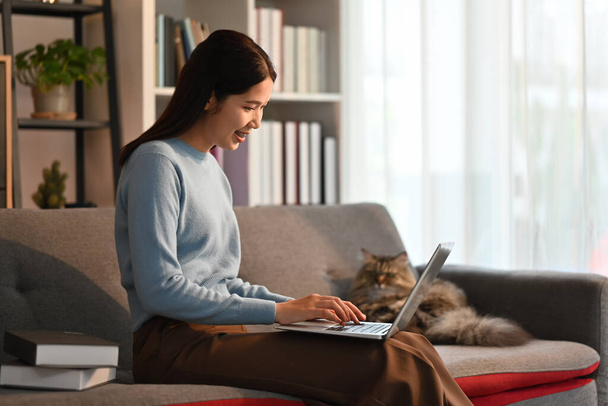 Leuke Aziatische vrouw met laptop computer op de bank en glimlachend moment met huiskat. Hoge kwaliteit foto - Foto, afbeelding