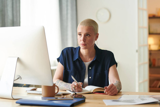 Seriöse Geschäftsfrau sitzt mit Computer am Tisch, blickt auf Monitor und macht sich während der Online-Konferenz Notizen im Notizblock - Foto, Bild