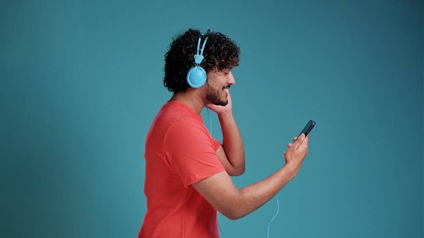 Cheerful handsome young indian guy in casual watching videos on smartphone, using new headset, looking at cell phone and smiling, in coral t-shirt on blue studio background, copy space. - Фото, зображення