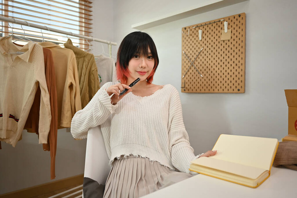 Νεαρή γυναίκα σχεδιάστρια μόδας που στέκεται κοντά σε σχάρα ρούχων, που εργάζονται σε νέα συλλογή γυναικείων ενδυμάτων στο σύγχρονο στούντιο. - Φωτογραφία, εικόνα
