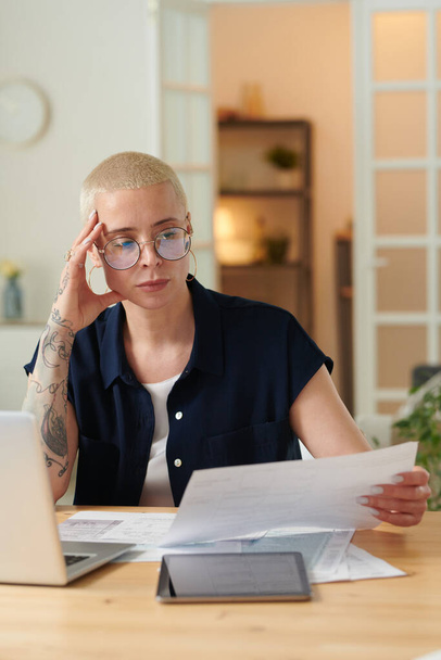 Vážná žena v brýlích zkoumá architektonický projekt na papíře pracující u stolu na notebooku - Fotografie, Obrázek