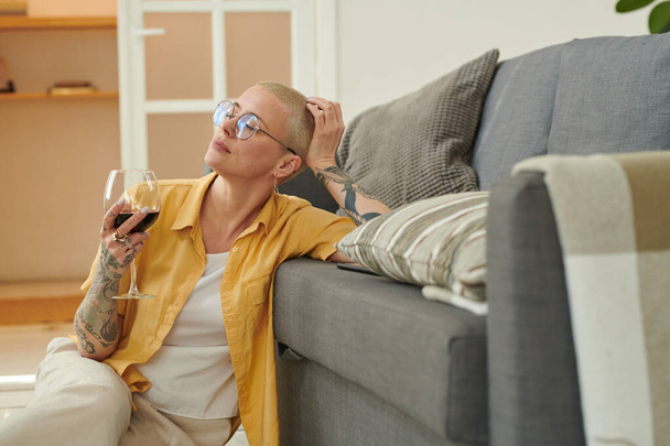 Красива жінка з коротким волоссям, сидячи на підлозі, спираючись на диван і насолоджуючись дозвіллям з червоним вином
 - Фото, зображення