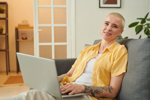 Dizüstü bilgisayardan satın alırken kameraya gülümseyen, kısa saçlı, olgun, zarif bir kadının portresi kanepede oturuyor. - Fotoğraf, Görsel