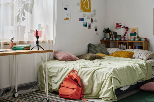Imagen horizontal de la habitación adolescente con acogedora cama y mesa con teléfono inteligente en trípode - Foto, imagen