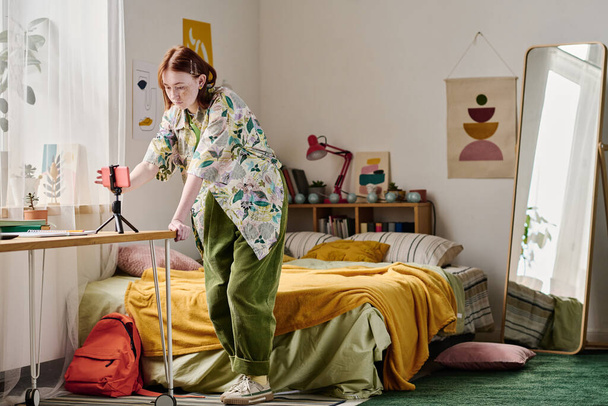 Chica adolescente encendiendo el teléfono inteligente de pie en el trípode para la transmisión en línea en su dormitorio - Foto, imagen