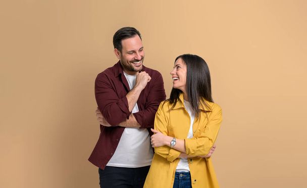 Happy romantic young couple with arms crossed smiling and looking at each other. Portrait of loving boyfriend and girlfriend standing together and posing joyfully over beige background - Photo, Image