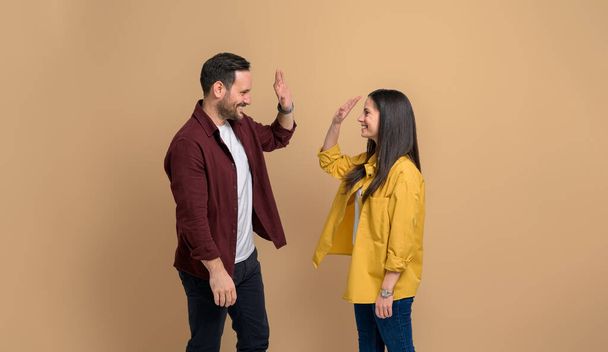 Smiling young couple in shirts giving high-five to each other and celebrating victory. Cheerful man and woman clapping hands and greeting while standing on isolated beige background - Photo, Image