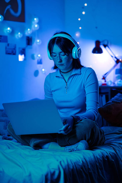 Chica adolescente en auriculares sentados en su cama y jugando videojuego en el ordenador portátil en la sala de neón oscuro - Foto, Imagen