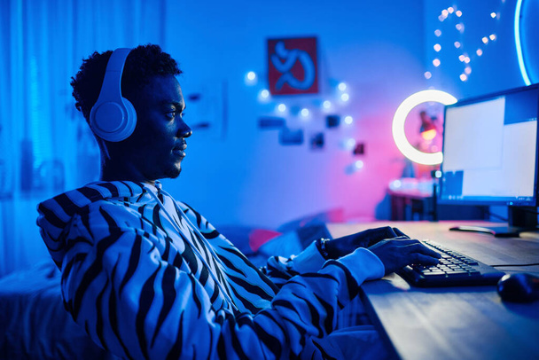Arican teenage boy in wireless headphones playing video game on computer while sitting at table in dark room - Photo, Image